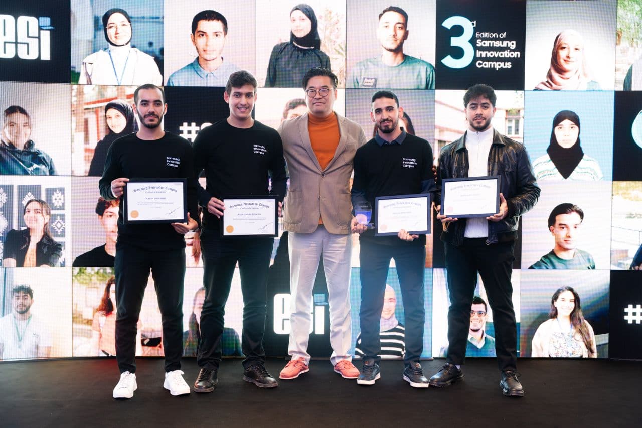 شامل وزملاؤه يحملون شهادات Samsung Innovation Campus أمام جدار من صور الطلاب